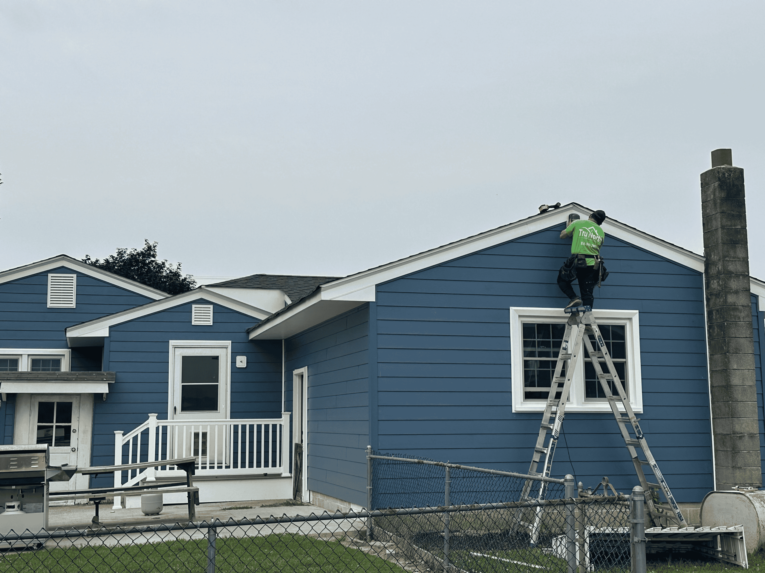 Home Exterior Painting Worker painting the blue house exterior with ladder and safety gear.